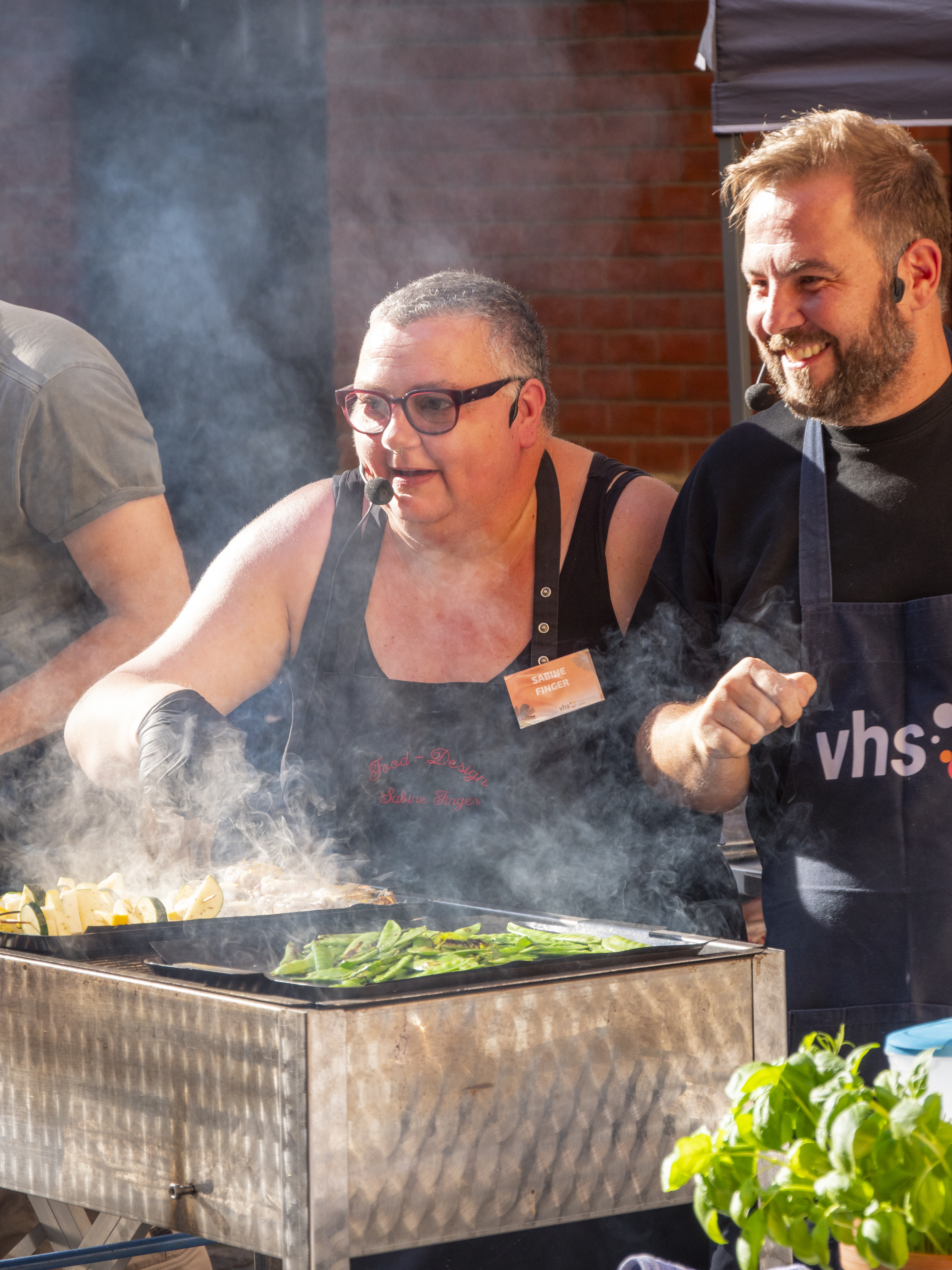 Live-Cooking Sommerfest 2024 eine Frau und ein Mann stehen am Grill und wenden Gemüse, beide sehen geschäftig und fröhlich aus