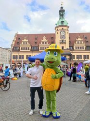 Ein Mann und eine Person in einem Schildkrötenkostüm stehen vor dem Alten Rathaus auf dem Leipziger Markt. Sie halten die Daumen in die Höhe. Die Schildkröte trägt einen gelben Anglerhut und blaue Flipflops. Sie hat grüne Haut udn einen orangefarbenen Panzer. Der Mann trägt ein weißes T-Shirt und eine schwarze Hose.