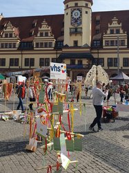 Auf dem Leipziger Marktplatz steht ein Baum aus Holz, an dem rote, grüne und gelbe Bänder hängen. Es hängen außerdem Karten aus grünem Papier mit weißer Schrift daran. Oben auf dem Baum ist ein Schild mit der Schrift "vhs Volkshochschule Leipzig" befestigt. Im Hintergrund sieht man das alte Rathaus.